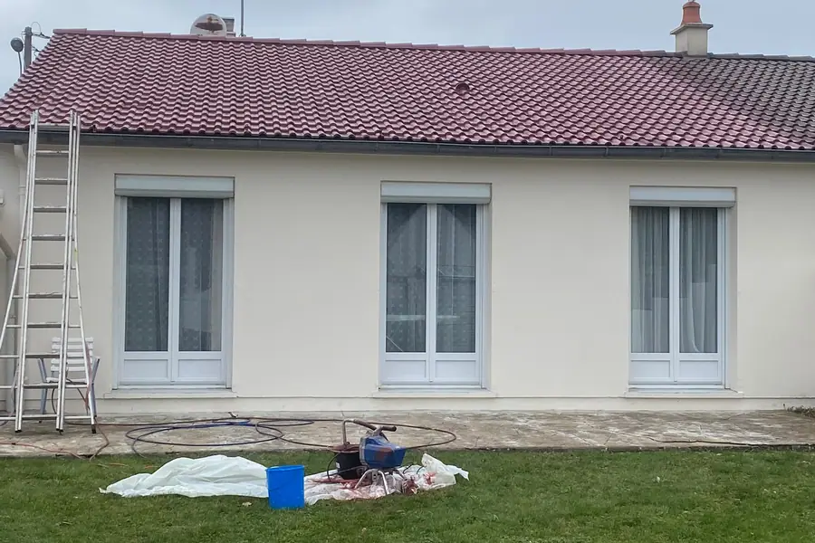 Mise en peinture d’une façade en cours : machine à projeter au pied du mur, bâche de protection au sol